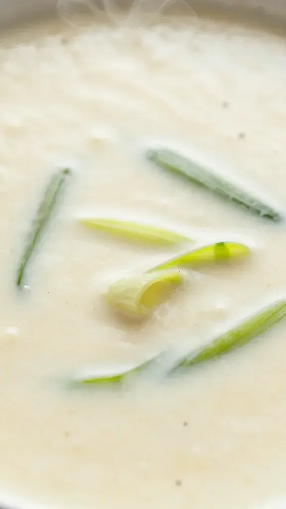 Close-up texture/detail shot: Close-up of the creamy, velvety soup surface with visible leek ribbons and a delicate pepper dusting, steamy and inviting, macro style