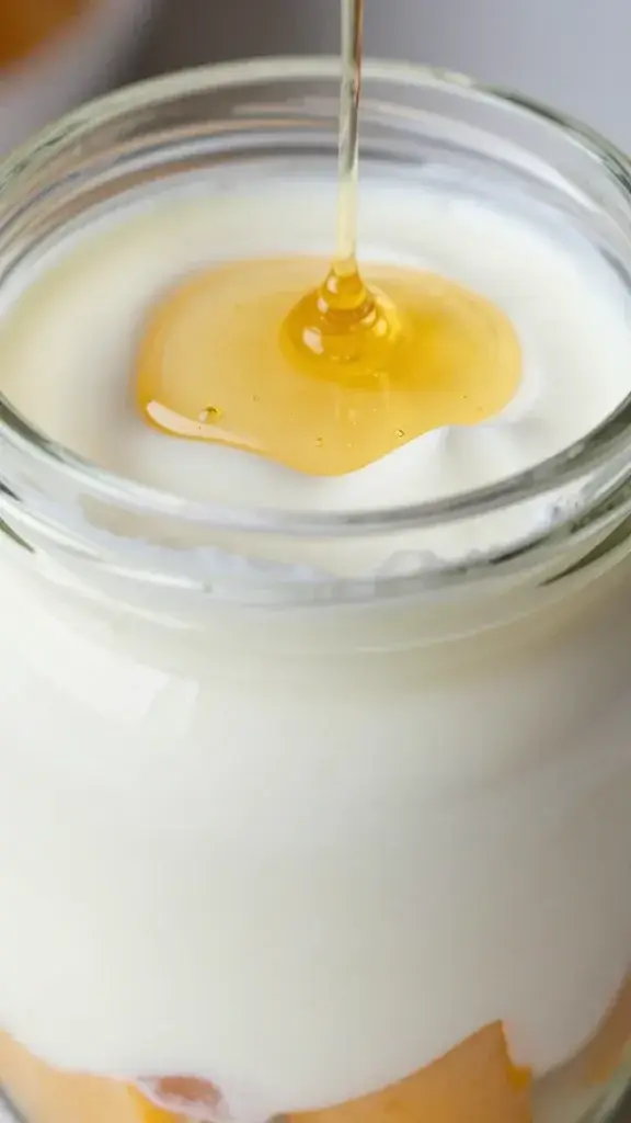 Close-up texture/detail shot: Creamy yogurt layers with visible fruit chunks in a glass jar, sizzling honey drizzle on top, droplets glistening, shallow depth of field, soft kitchen light.